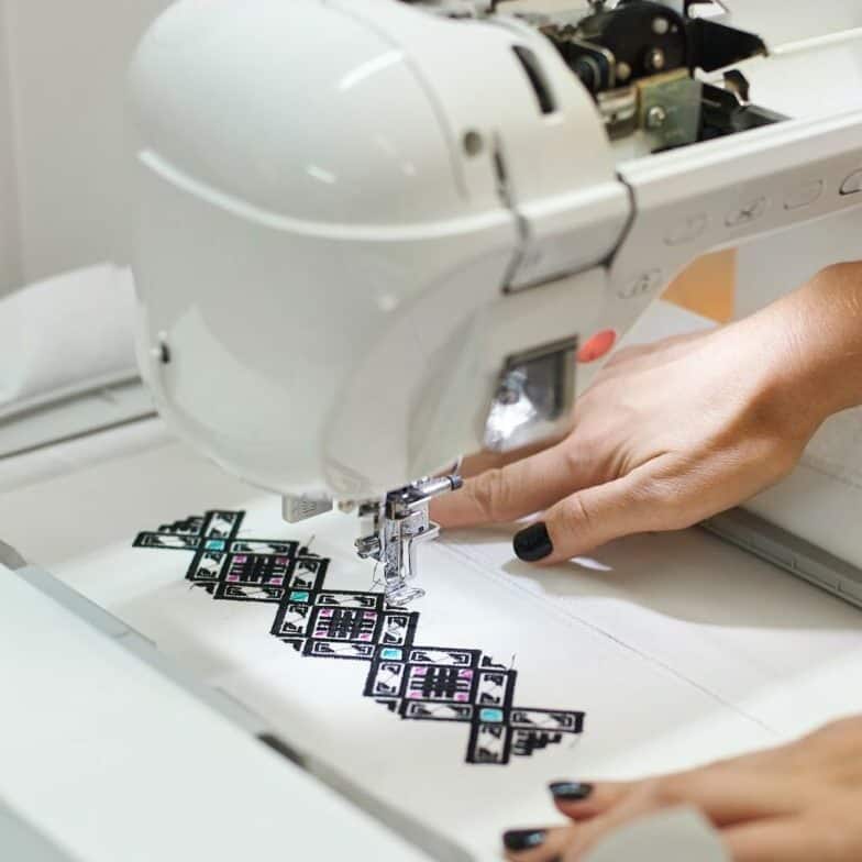 Close-up of sewing machine creating colorful abstract geometrical pattern on white fabric.